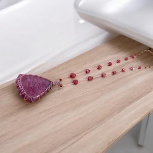 Puede incluir: Un collar hecho a mano con un gran colgante de piedra preciosa rosa de forma irregular. El colgante está envuelto en alambre y cuelga de una delicada cadena con cuentas rosas a juego. El collar descansa sobre una superficie de madera clara.