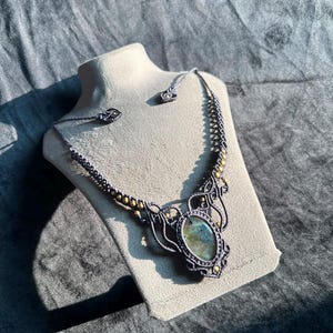 Puede incluir: Un collar de macramé con una piedra labradorita ovalada central con destellos iridiscentes azules y verdes. El collar está elaborado con cordón gris oscuro con patrones de nudos intrincados y pequeñas cuentas metálicas doradas.