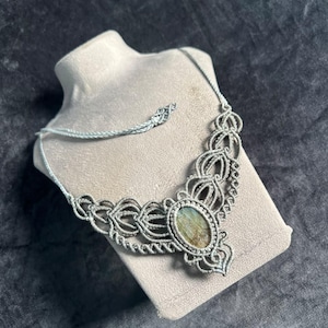 May include: A macrame necklace featuring a central oval-shaped stone with iridescent green and gold tones. The stone is encased in intricate grey cord knotwork, forming a decorative pattern that extends along the sides of the necklace cord.