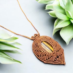May include: A handcrafted macrame necklace featuring a brown pendant with an oval tiger's eye stone. The pendant has intricate woven patterns and hangs from a matching cord, showcasing a bohemian style.