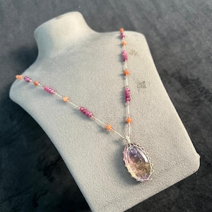 Puede incluir: Un collar con colgante de piedras preciosas con una piedra grande, ovalada y multicolor. El colgante está envuelto en alambre y cuelga de una delicada cadena adornada con cuentas rosas y naranjas. El collar se exhibe en un busto gris.