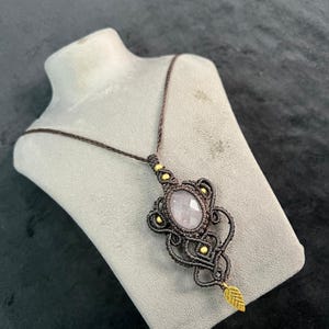 May include: A handmade macramé necklace with a light purple oval gemstone pendant. The pendant is framed by intricate dark brown macramé knots and accented with small gold-coloured beads. A small gold leaf charm hangs below.