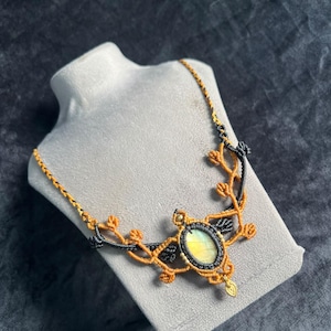 Può includere: Una collana in macramè con una pietra di labradorite ovale centrale caratterizzata da riflessi iridescenti gialli e blu. Il ciondolo è incorniciato da intricati motivi a cordoncino annodato arancione e nero che formano dei rami.