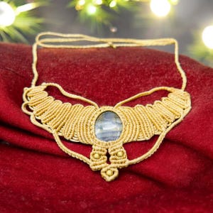 May include: A handcrafted macrame necklace with a central oval-shaped stone. The necklace features intricate woven patterns in a light beige color, with a red background. The stone has a blue and gray color.