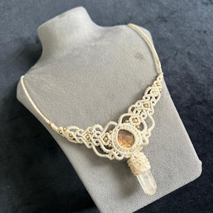 May include: Cream macrame necklace with a central, round, brown stone and a clear crystal pendant. The necklace features intricate knotting and small gold-coloured beads. Displayed on a grey bust against a dark background.