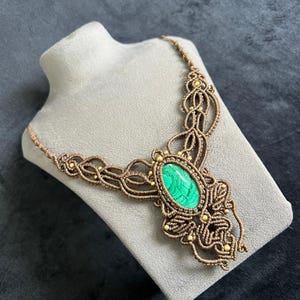 May include: A handmade macramé necklace with a large, oval green stone centrepiece. The necklace is crafted from brown cord and embellished with small gold beads. The intricate knotting creates a detailed, bohemian design, suitable for any occasion.