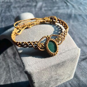 May include: A macrame bracelet crafted from tan cord featuring an oval-shaped dark green stone at the center. The design includes intricate knotted patterns and small metallic beads integrated into the structure, with an adjustable cord closure.