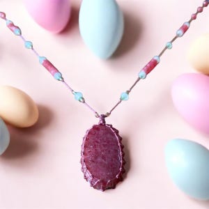 May include: A handcrafted necklace with a large, oval pendant in burgundy and pink hues. The pendant hangs from a delicate cord embellished with small, light blue and pink beads. Pastel-coloured egg-shaped decorations are in the background.