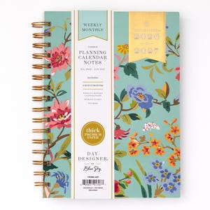 May include: A spiral-bound planner with a floral design in shades of pink, blue, and orange on a teal background. The planner includes the text "Weekly Monthly", "Planning Calendar Notes", and "Day Designer" for the years 2026-2027.