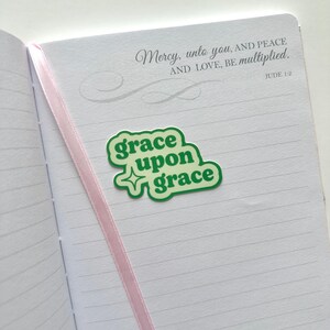 May include: An open notebook with lined pages and a pink ribbon bookmark. A green sticker with the words "grace upon grace" is placed on the page. The text "Mercy, unto you, AND PEACE AND LOVE, BE multiplied. JUDE 1:2" is printed at the top.