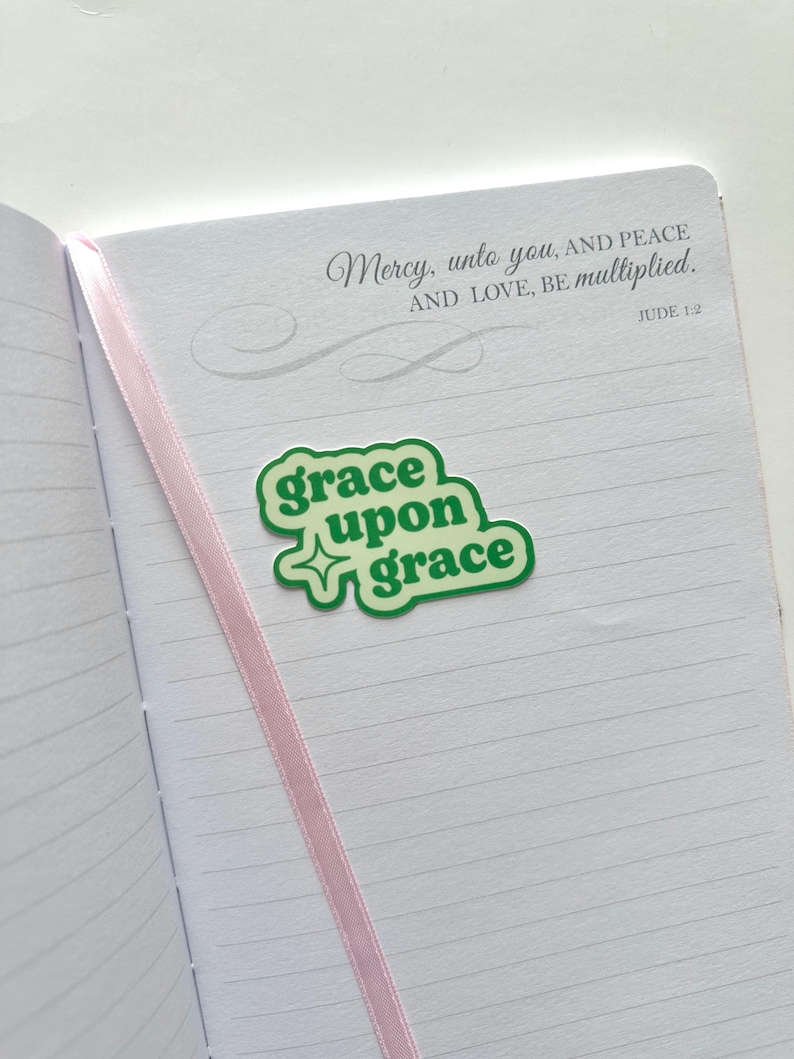 May include: An open notebook with lined pages and a pink ribbon bookmark. A green sticker with the words "grace upon grace" is placed on the page. The text "Mercy, unto you, AND PEACE AND LOVE, BE multiplied. JUDE 1:2" is printed at the top.