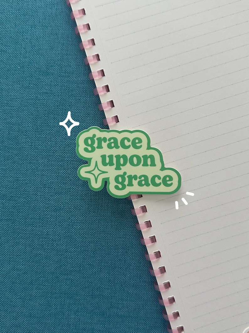 May include: A green sticker with the words "grace upon grace" in a stylized font. The sticker is on a white lined notebook with a pink spiral binding, set against a teal background.