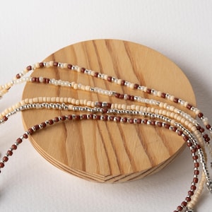 May include: Close-up of several beaded necklaces in shades of brown, cream, and silver. The necklaces are displayed on a small, round, wooden stand. The beads are small and evenly spaced, creating a delicate and intricate design.