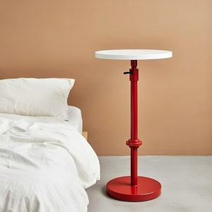 May include: A red adjustable side table with a white circular top. The table has a round base and a central pole that can be adjusted in height. The table is next to a bed with white bedding.