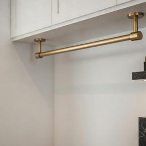 May include: A gold-toned metal closet rod suspended from a white ceiling. The rod is cylindrical and supported by two vertical posts. The design is minimalist and modern, suitable for a contemporary closet or storage space.