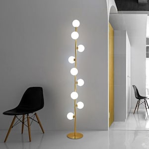 May include: A modern floor lamp with a gold-colored stem and a series of white globe lights. The lamp stands tall, with a round base. The background features a gray wall, a black chair, and a white floor.