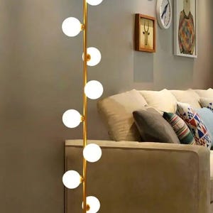 May include: A modern floor lamp with a gold-colored metal frame and multiple spherical white light bulbs. The lamp has a round base and a vertical design, illuminating a living room setting. The lamp is a decorative lighting fixture.