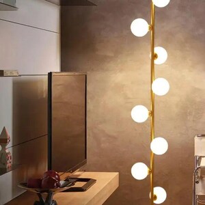 May include: A modern floor lamp with a gold-colored stem and multiple spherical, white glass light bulbs. The lamp is positioned in a room with a neutral-toned wall and wooden furniture. The lamp's design is minimalist and contemporary.