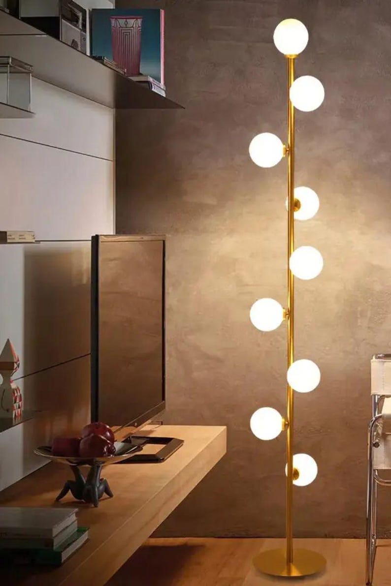 May include: A modern floor lamp with a gold-colored stem and multiple spherical, white glass light bulbs. The lamp is positioned in a room with a neutral-toned wall and wooden furniture. The lamp's design is minimalist and contemporary.