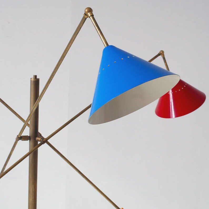Może przedstawiać: Lampa podłogowa w stylu mid-century modern z mosiężnym wykończeniem i dwoma stożkowymi kloszami. Jeden klosz jest w kolorze jasnoniebieskim, a drugi czerwonym. Lampa ma minimalistyczny design z regulowanymi ramionami i solidną podstawą.