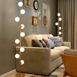 May include: A modern floor lamp with a gold finish and multiple spherical white lights. The lamp stands tall next to a beige sofa with decorative pillows. The room has artwork and a neutral color scheme.