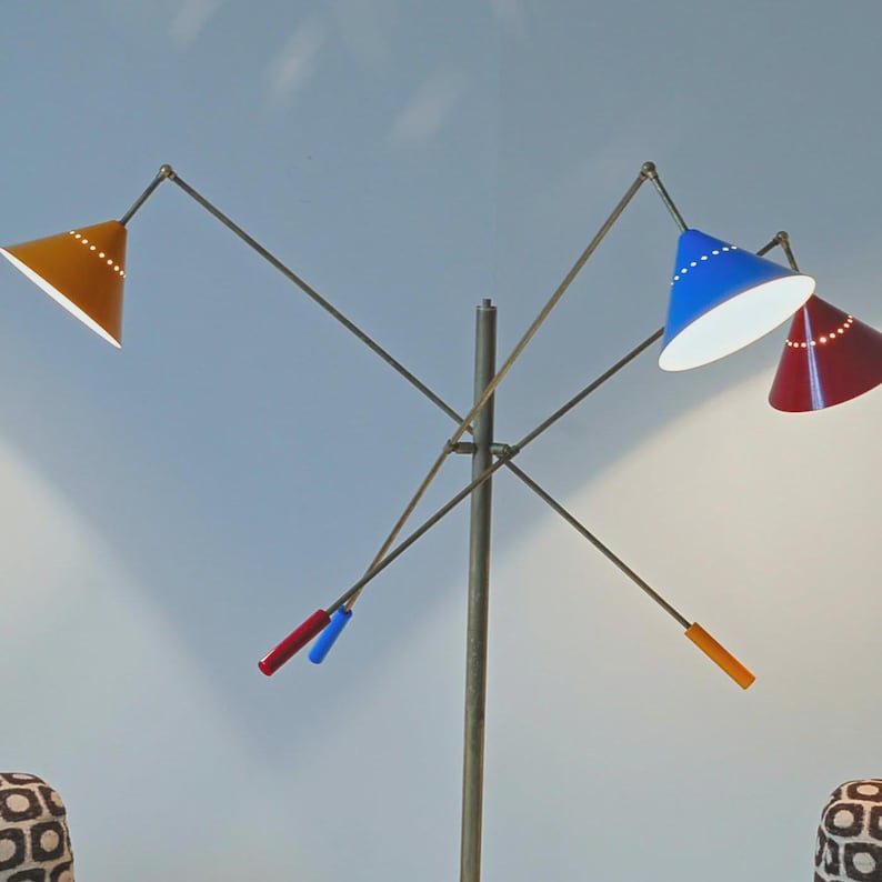 Może przedstawiać: Wielokolorowa lampa podłogowa o nowoczesnym designie z połowy wieku. Lampa posiada trzy stożkowe klosze w kolorze ż&oacute;łtym, niebieskim i czerwonym, każdy z rzędem małych otwor&oacute;w. Ramiona lampy są regulowane i rozciągają się z centralnego słupa.