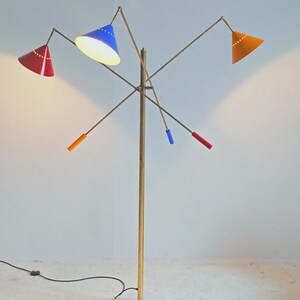 Może przedstawiać: Lampa podłogowa w stylu retro z mosiężnym trzonem i wieloma ramionami, z kt&oacute;rych każde ma stożkowy klosz. Klosze są czerwone, niebieskie i pomarańczowe. Design lampy nawiązuje do nowoczesnych styl&oacute;w z połowy wieku.
