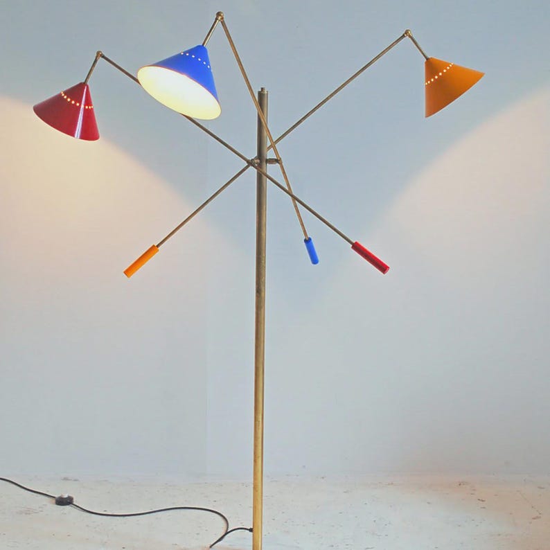 Może przedstawiać: Lampa podłogowa w stylu retro z mosiężnym trzonem i wieloma ramionami, z kt&oacute;rych każde ma stożkowy klosz. Klosze są czerwone, niebieskie i pomarańczowe. Design lampy nawiązuje do nowoczesnych styl&oacute;w z połowy wieku.