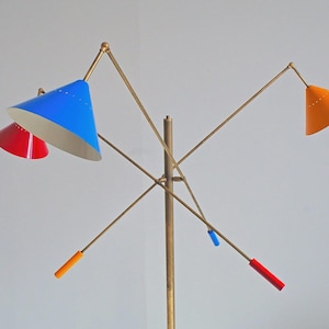 Może przedstawiać: Lampa podłogowa w stylu mid-century modern z mosiężnym wykończeniem i wieloma ramionami. Lampa posiada stożkowe klosze w kolorze czerwonym, niebieskim i pomarańczowym. Ramiona mają kolorowe akcenty w kolorze niebieskim, pomarańczowym i czerwonym. Stylowa lampa.