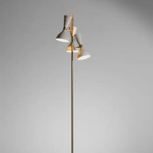 Op de afbeelding: Een vloerlamp uit het midden van de eeuw met een messingkleurige paal en vier armaturen. De lampenkappen zijn lichtgrijs van kleur. De lamp is aangesloten op een zwart stopcontact.