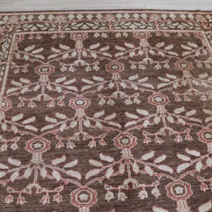 May include: A rectangular area rug featuring a dark brown background with a repeating floral and vine pattern in shades of cream and muted pink. The design includes stylized flowers and leaves arranged in a grid, framed by a decorative border.