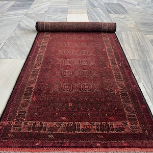 May include: A rolled-up, rectangular, red rug with a detailed geometric pattern. The rug has a dark border and a central field with repeating square motifs. The rug is partially unrolled, revealing its intricate design and texture.