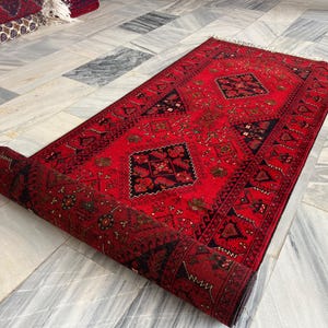 Puede incluir: Una alfombra rectangular con un fondo rojo vibrante y patrones geométricos intrincados en negro y azul oscuro, que incluyen motivos de diamantes centrales y bordes detallados, con una parte de la alfombra enrollada.