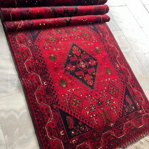 Op de afbeelding: Een rechthoekig vloerkleed met een levendige rode achtergrond en ingewikkelde zwarte en donkerblauwe geometrische patronen. Het kleed heeft een centraal ruitmotief omringd door gestileerde florale en tribale elementen, afgewerkt met witte franjes.