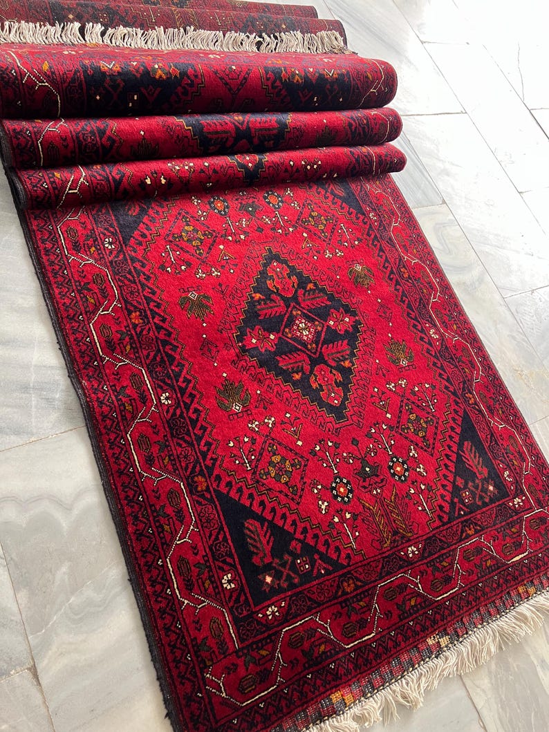 Op de afbeelding: Een rechthoekig vloerkleed met een levendige rode achtergrond en ingewikkelde zwarte en donkerblauwe geometrische patronen. Het kleed heeft een centraal ruitmotief omringd door gestileerde florale en tribale elementen, afgewerkt met witte franjes.