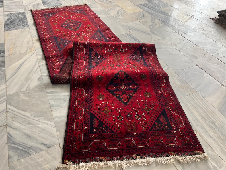 Op de afbeelding: Een rechthoekige loper met een dieprode achtergrond, voorzien van donkerblauwe geometrische ruitpatronen en ingewikkelde bloemmotieven. Het kleed heeft een brede rand met herhalende lineaire ontwerpen en korte witte franjes aan de uiteinden.