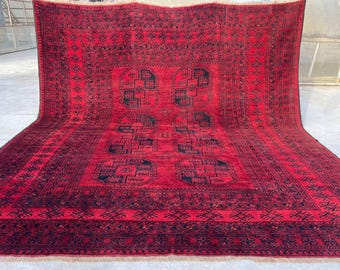 Op de afbeelding: Een groot, rechthoekig rood tapijt met ingewikkelde zwarte geometrische patronen. Het tapijt heeft een centraal ontwerp met vier zich herhalende vierkante motieven en een gedetailleerde rand. Het tapijt wordt gepresenteerd op een lichtgekleurd oppervlak.
