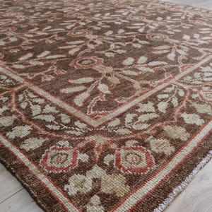 May include: A rectangular area rug featuring a deep brown background with a repeating floral and vine pattern in cream and muted coral tones. The design includes a prominent border with stylized blossoms and leaves framing the central field.