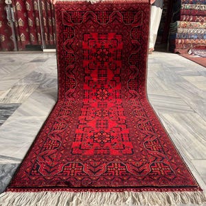 May include: A vibrant red and black oriental rug with intricate geometric patterns. The rug features a central medallion design and a detailed border. The rug has a fringe on both ends and is displayed on a marble floor.