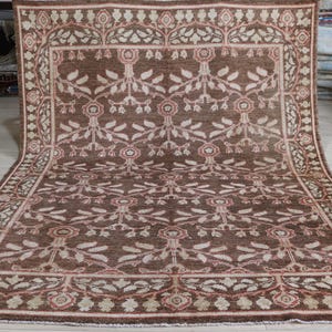 May include: A rectangular area rug featuring a deep brown background with a repeating floral and vine pattern in shades of cream, beige, and muted pink. The rug has a wide decorative border with a similar botanical motif and a fringed edge.