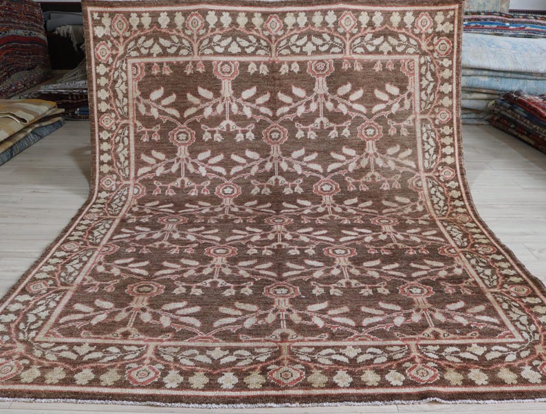 May include: A rectangular area rug featuring a deep brown background with a repeating floral and vine pattern in shades of cream, beige, and muted pink. The rug has a wide decorative border with a similar botanical motif and a fringed edge.