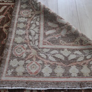 May include: A woven rug featuring a brown background with a cream and coral floral pattern. The rug is folded at the corner, revealing the underside of the fabric. The design consists of repeating botanical motifs and geometric border elements.