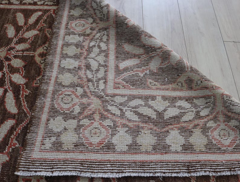 May include: A woven rug featuring a brown background with a cream and coral floral pattern. The rug is folded at the corner, revealing the underside of the fabric. The design consists of repeating botanical motifs and geometric border elements.