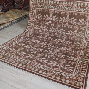 May include: A large rectangular area rug featuring a deep brown background with a repeating floral and vine pattern in cream and muted red. The rug has a wide decorative border with a similar botanical motif and a fringe along the edges.