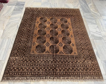 Op de afbeelding: Een rechthoekig vloerkleed met een centraal bruinachtig beige veld met herhalende donkerbruine achthoekige motieven en kleine geometrische symbolen. De brede rand toont ingewikkelde donkerbruine patronen en de randen zijn afgewerkt met korte franjes.