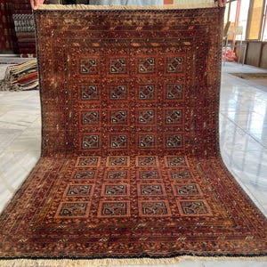 May include: A rectangular area rug featuring a deep red and brown color palette with a repeating grid of square motifs. The rug displays intricate geometric patterns within each square, a wide decorative border, and off-white fringe at the ends.