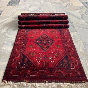 Op de afbeelding: Een rechthoekig vloerkleed met een dieprode achtergrond, een centraal donkerblauw ruitvormig medaillon en ingewikkelde geometrische patronen in zwart, goud en wit. Het kleed heeft een dik opgerold bovendeel en witte franjes aan de onderkant.