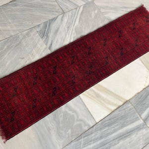 May include: A rectangular, red runner rug with a repeating geometric pattern in black. The rug has a fringed edge and is displayed on a marble tile surface. This rug is suitable for hallways or entryways.