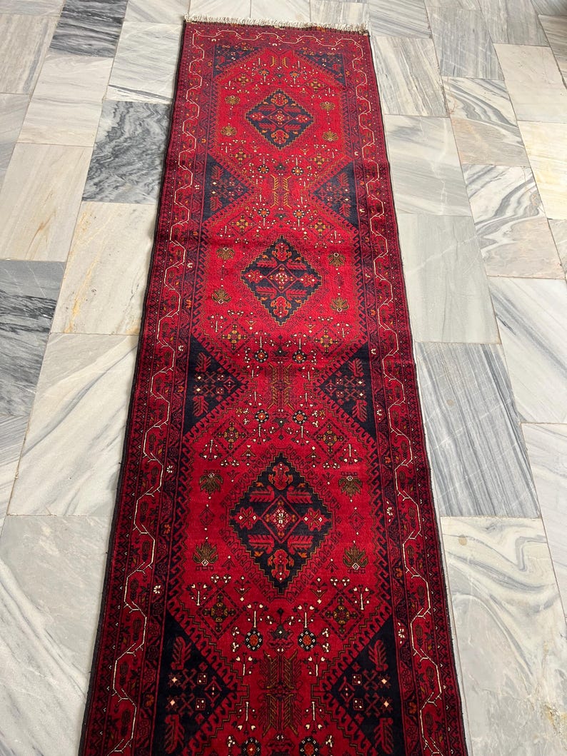 Op de afbeelding: Een lang, rechthoekig loperkleed met een dieprode achtergrond en drie opvallende donkerblauwe ruitvormige medaillons. Het kleed is versierd met ingewikkelde geometrische patronen en kleine bloemmotieven in zwart, wit en goud, afgewerkt met korte franjes.
