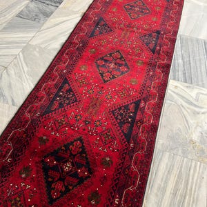 Op de afbeelding: Een lange, smalle loper met een levendige rode achtergrond en herhalende donkerblauwe geometrische ruitmotieven. Het kleed is versierd met ingewikkelde, kleinschalige bloem- en tribale patronen in tinten goud, wit en zwart.